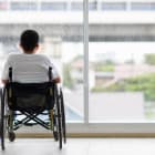 Back view of young Asian handicapped teenage boy without legs sitting on wheel chair and looking to outside of home with copy space on right.
