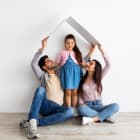 Housing for young family concept. Young eastern father, mother and daughter under symbolic roof over light wall