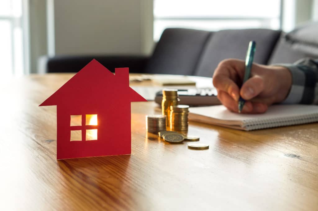 Main d'homme écrivant sur un cahier avec devant une pile de pièce et un photophore rouge en forme de maison